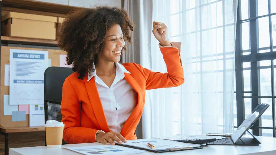 A jubilant businesswoman in a bright orange blazer celebrates a success at her desk, with a laptop open and financial reports spread out in front of her, in a sunny office space with charts displayed on the wall behind, signifying professional achievement.