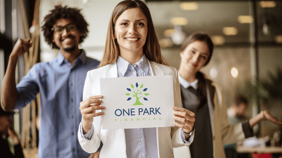 A smiling woman holding a sign for "One Park Financial" stands in the foreground, with her enthusiastic team, including a man giving a thumbs up, in the background.