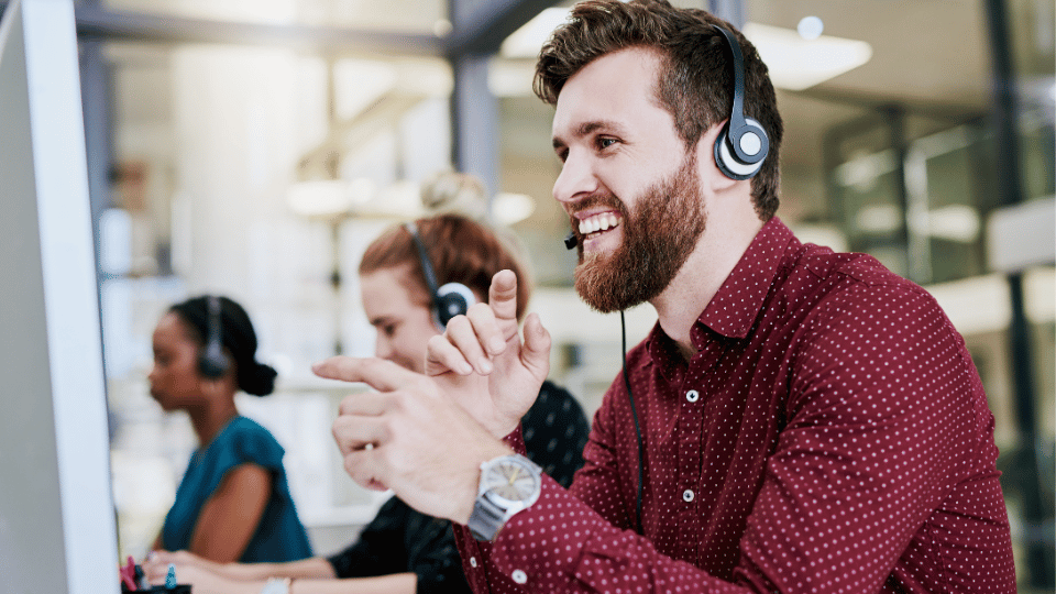 A customer service representative with headphones gesturing with his hand while smiling, with colleagues in the background, representing a positive customer support experience.