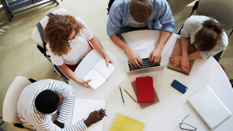 Overhead view of a diverse team working together at a round table, with one person typing on a laptop and others writing notes, symbolizing collaborative team effort and diversity in the workplace.