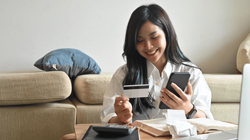 A smiling woman with a credit card and phone handles finances on a sofa, with bills, a calculator, and a laptop on the table in front of her.