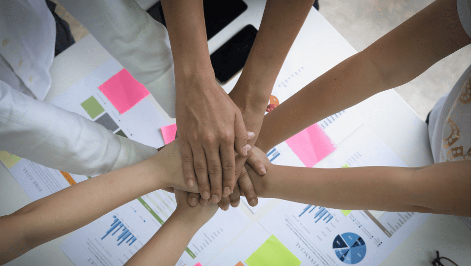 "Overhead view of a diverse group of people's hands coming together in a unified stack over a table covered with financial charts and business reports, symbolizing teamwork and collaboration in a business or financial planning setting