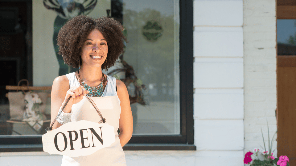 A joyful woman business owner displays an 'OPEN' sign, representing success enabled by small business loans for women.