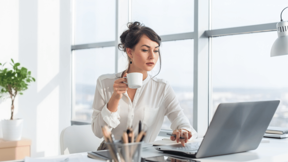 A woman at a desk with a laptop, holding a coffee mug, with large windows in the background.