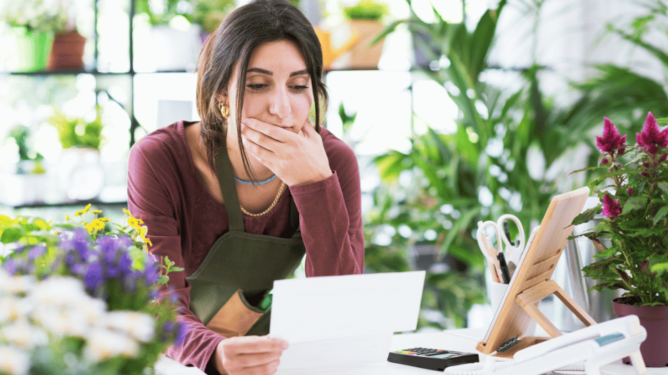 A concerned woman in a garden shop reads a document, possibly troubled by strict bank loan criteria for her business.