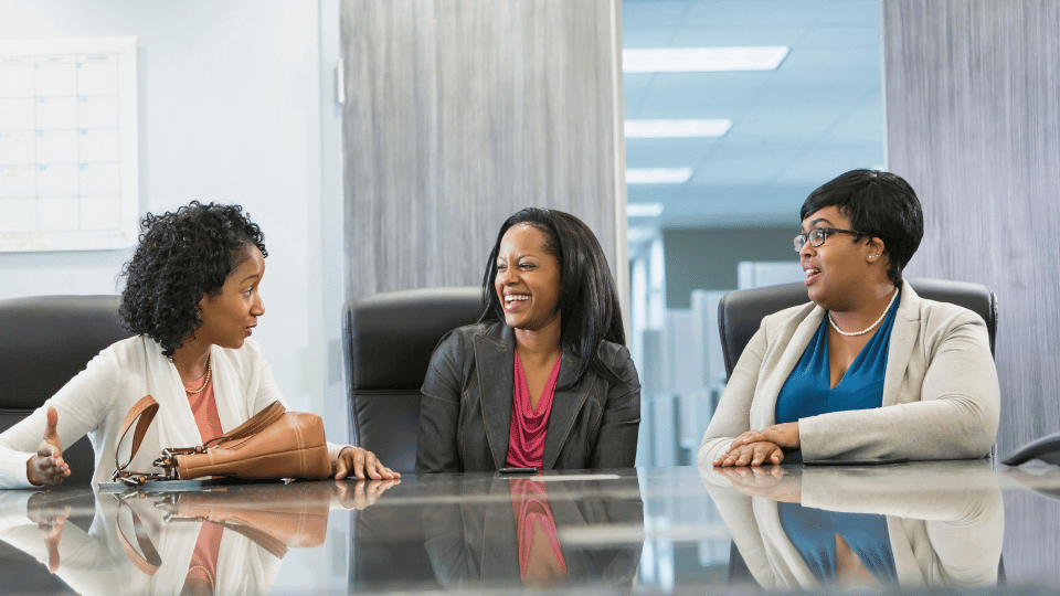 Three women, possibly colleagues, are sitting around a conference table in an office, engaged in a friendly and animated conversation. They are dressed in business casual attire, suggesting a professional setting. The office has a modern decor with a bright and open atmosphere.